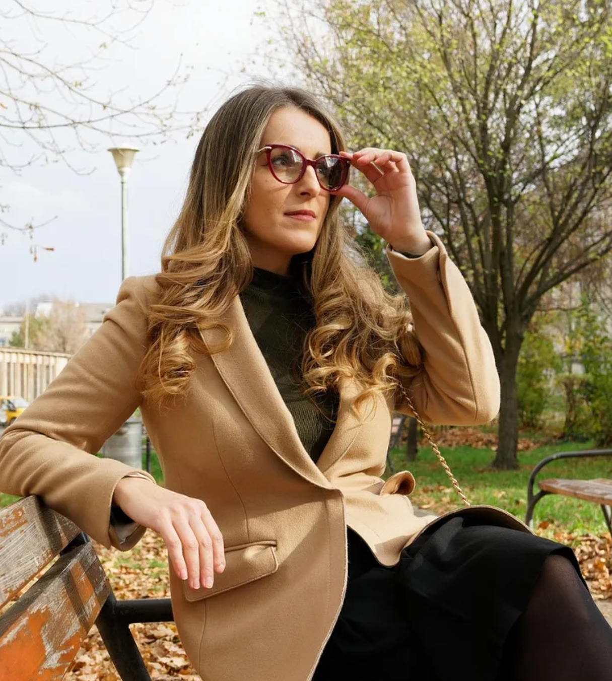a woman sitting on a park bench wearing brown blazer and shades